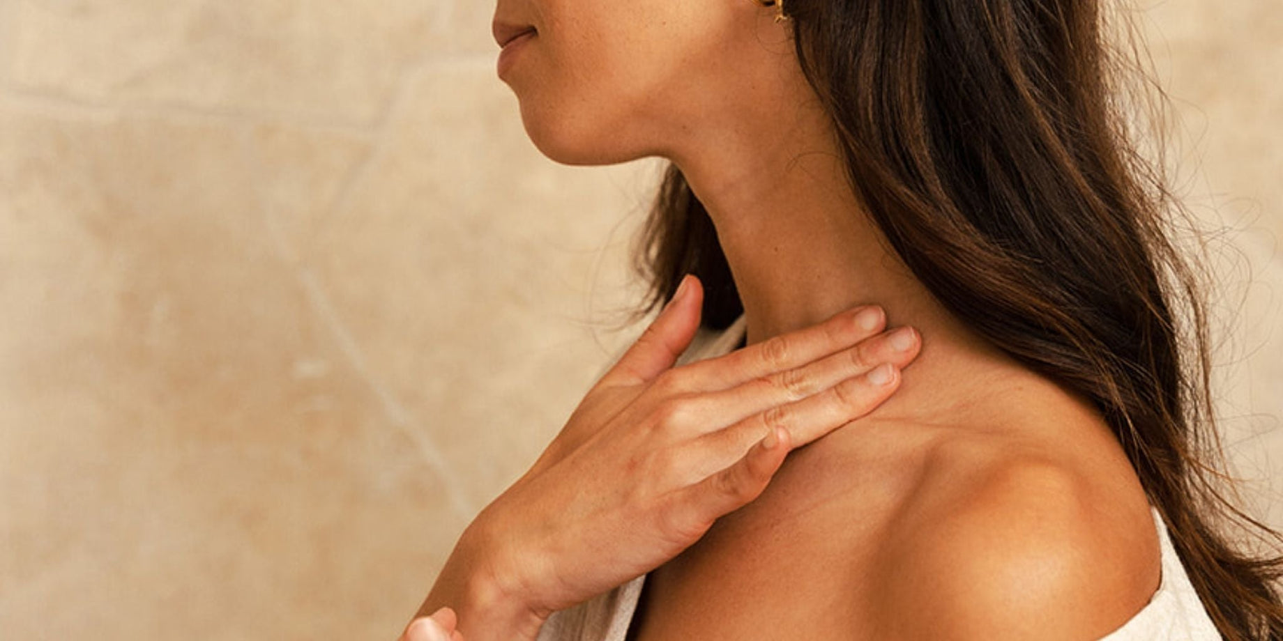 A woman with healthy, balanced skin touching her neck and shoulder against a warm limestone wall — the result of a consistent Certified Organic skincare routine.