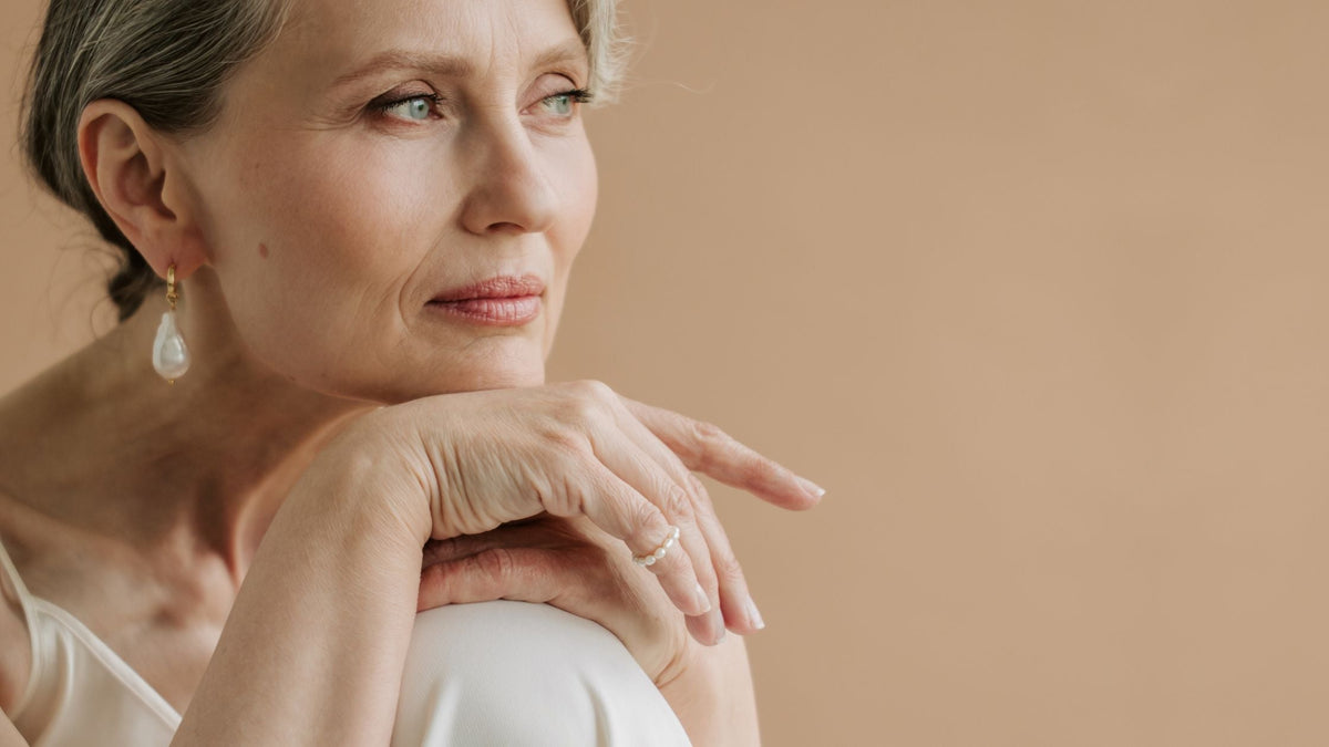 A woman with silver hair and a pearl drop earring resting her chin on her hand, looking into the distance against a warm beige background.