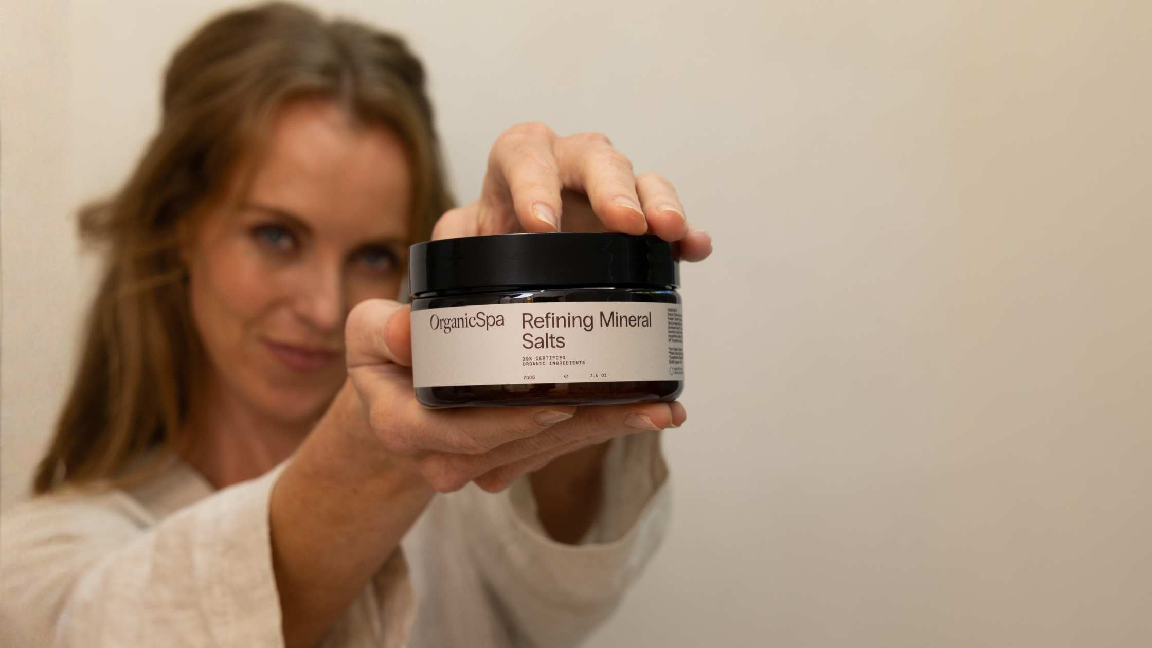 Woman holding a jar of OrganicSpa Refining Mineral Salts towards the camera, with a soft neutral background.