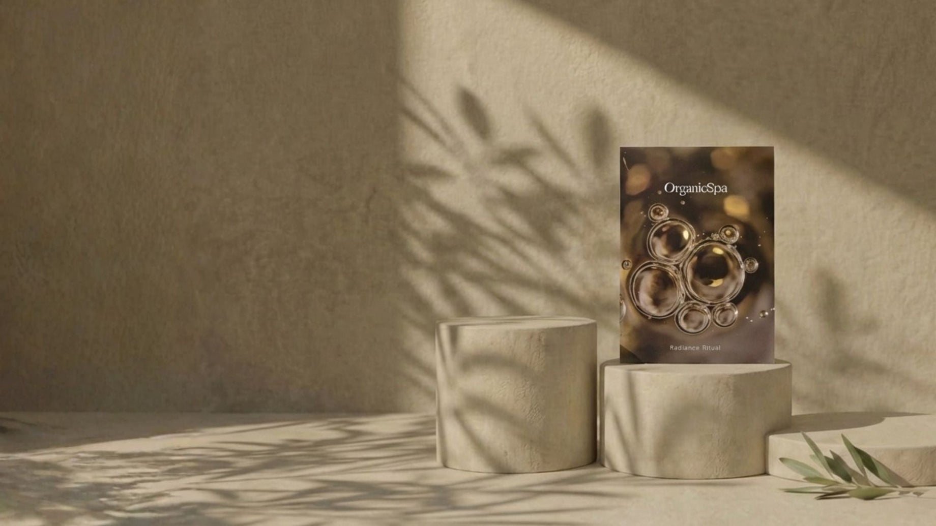 OrganicSpa Radiance Ritual box standing on stone cylindrical plinths with dramatic olive branch shadow play across a warm sandy textured wall