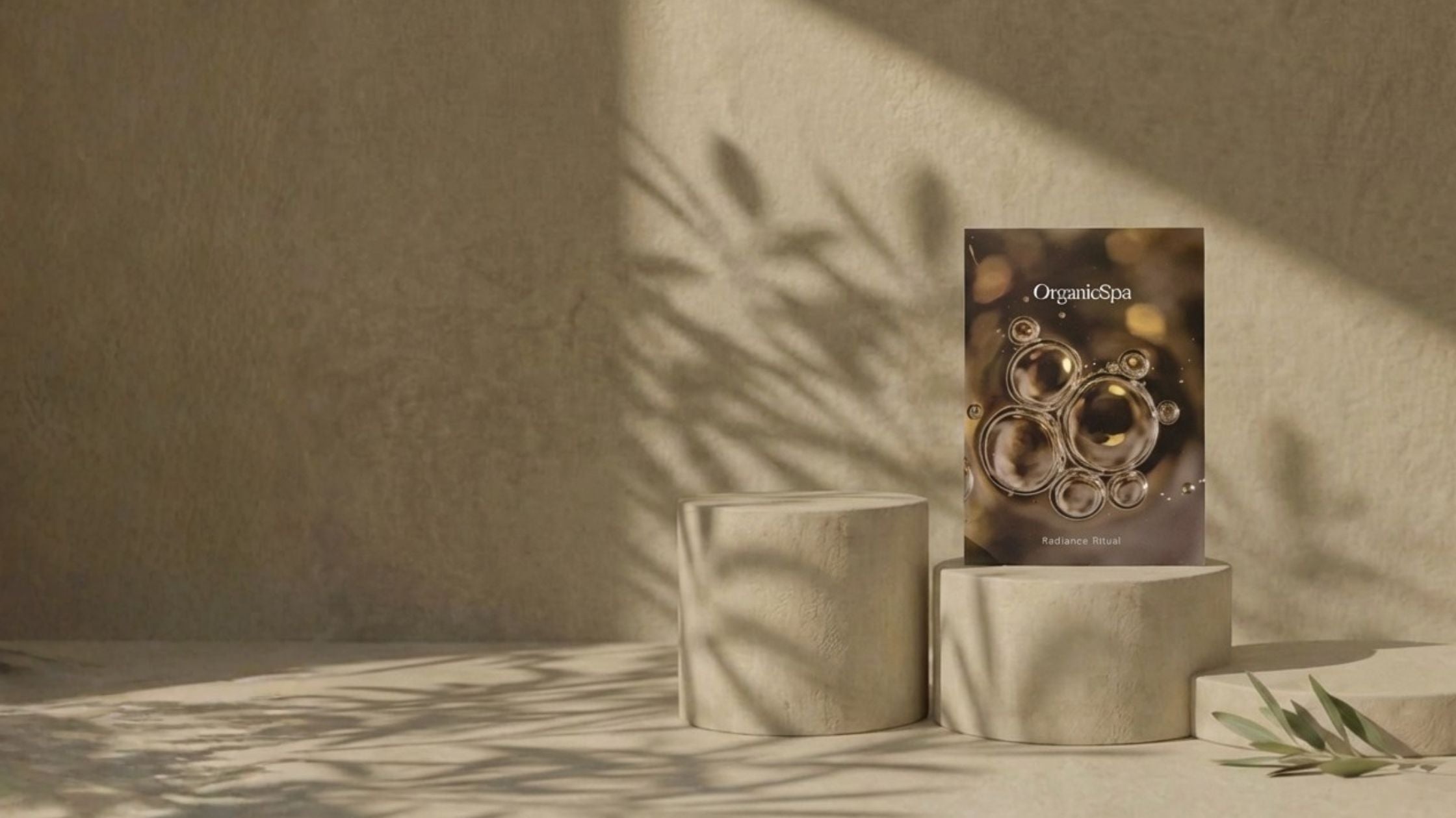 OrganicSpa Radiance Ritual box standing on stone cylindrical plinths with dramatic olive branch shadow play across a warm sandy textured wall