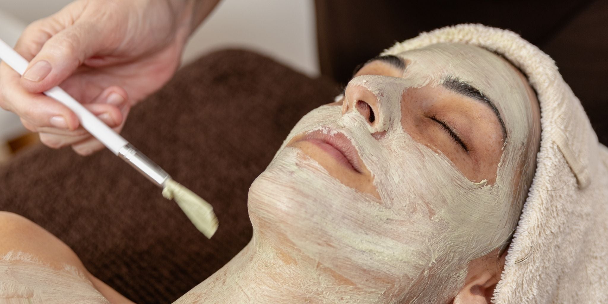 A skin therapist applying a clay mask to a client's face using a treatment brush.A skin therapist applying a clay mask to a client's face using a treatment brush. The client is reclined with eyes closed, hair wrapped in a white towel, draped with a chocolate brown treatment cloth.