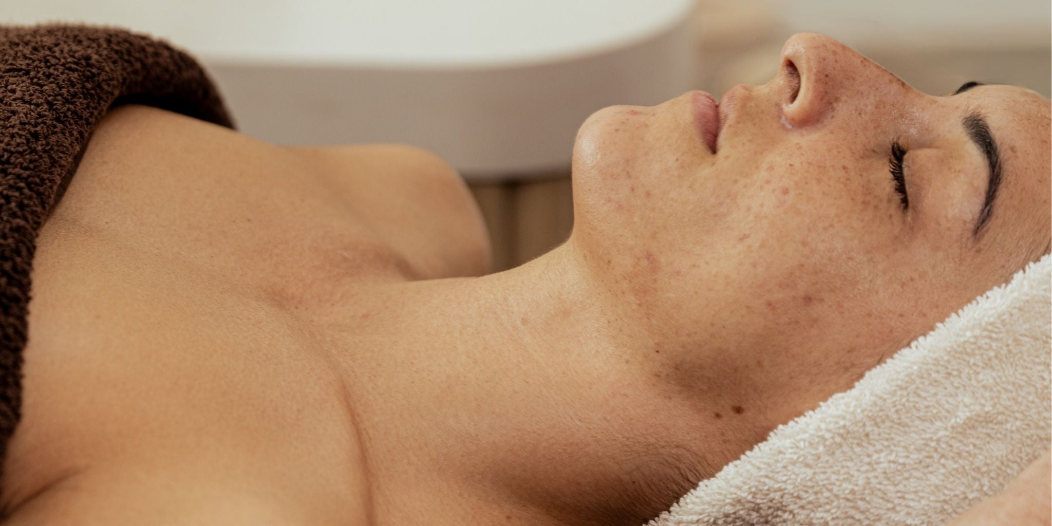 A close profile shot of a client reclined on a treatment table, eyes closed, skin bare and relaxed.A close profile shot of a client reclined on a treatment table, eyes closed, skin bare and relaxed. Draped in chocolate brown and white towels, with a clean white basin visible in the background.