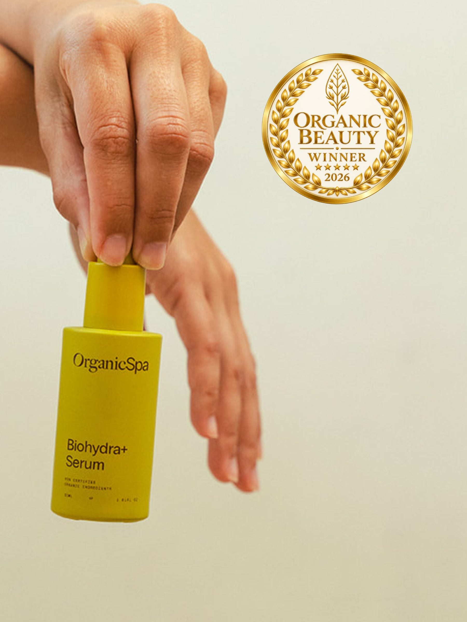OrganicSpa Biohydra+ Serum, a chartreuse bottle held upright in one hand against a pale background. Gold laurel badge beside it: Organic Beauty winner 2026.