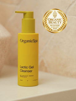 OrganicSpa Lactic Gel Cleanser, a chartreuse 140ml pump bottle marked 80% certified organic ingredients, on a cream ledge. Gold laurel badge beside it: Organic Beauty winner 2026.