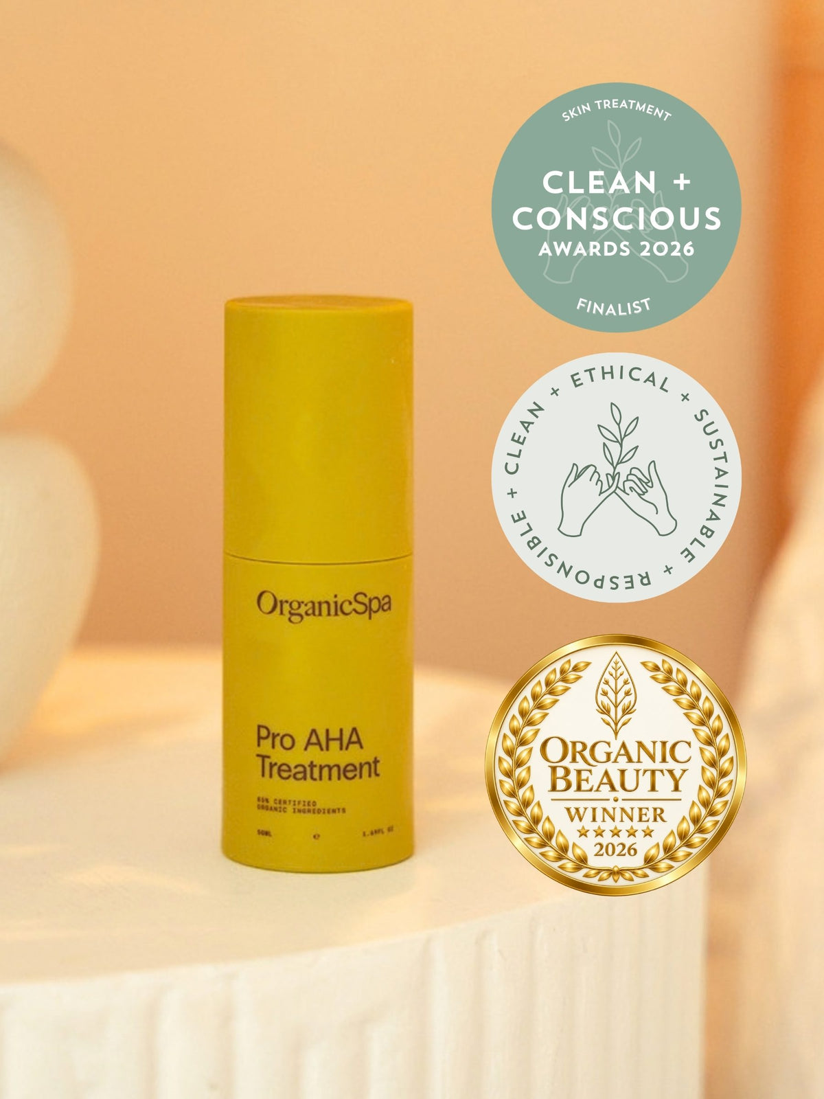 OrganicSpa Pro AHA Treatment, a chartreuse bottle on a cream plinth. Three badges beside it: finalist in the Skin Treatment category at the Clean + Conscious Awards 2026, the awards' clean, ethical, sustainable and responsible seal, and Organic Beauty winner 2026.