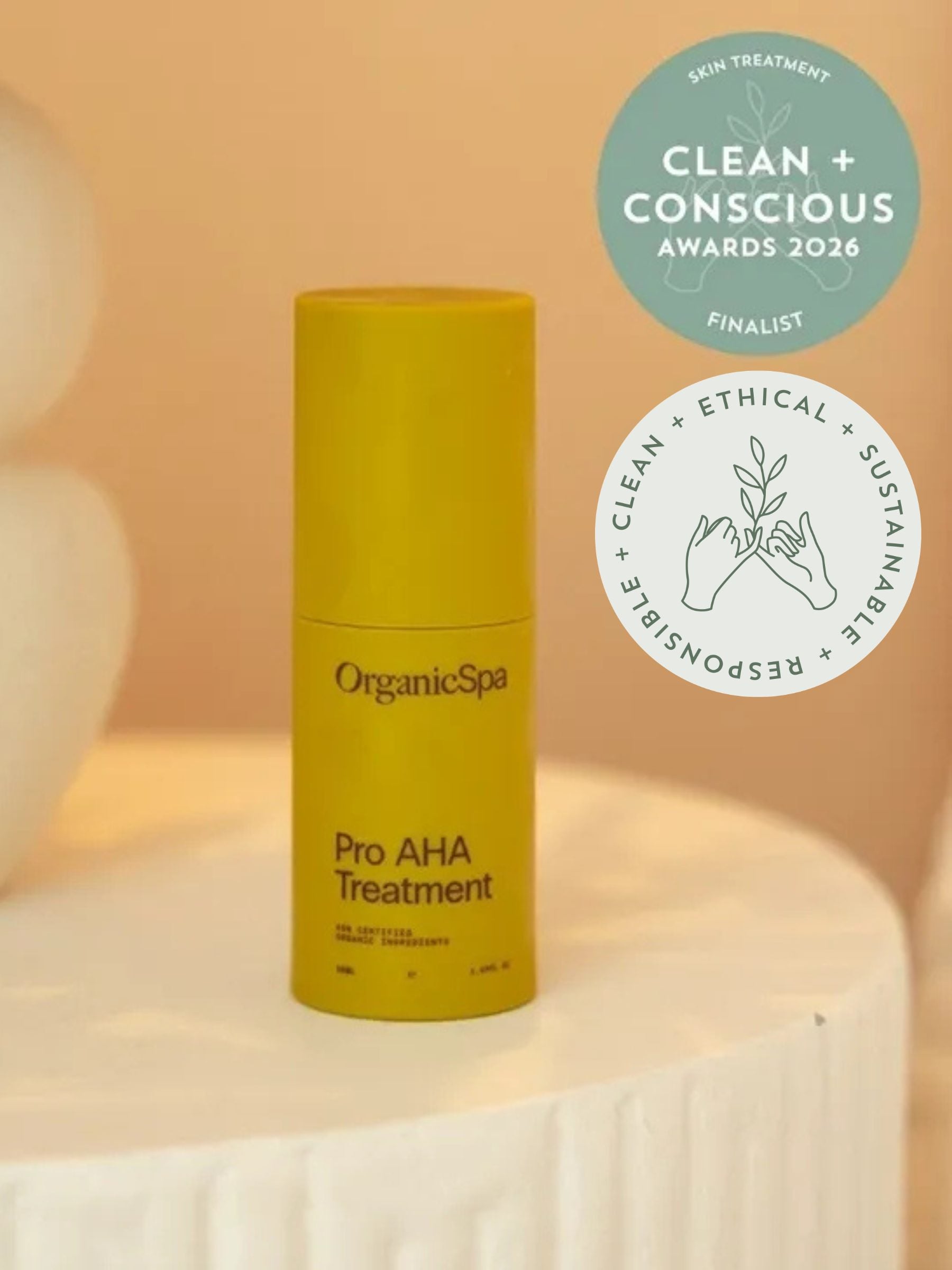 OrganicSpa Pro AHA Treatment, a chartreuse bottle on a cream plinth. Award badges beside it: finalist in the Skin Treatment category at the Clean + Conscious Awards 2026, and the awards' clean, ethical, sustainable and responsible seal.
