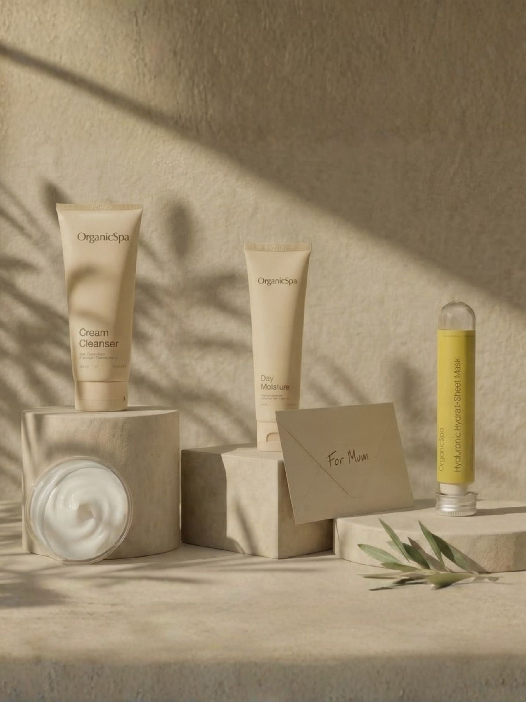OrganicSpa Mother's Day Radiance Ritual gift collection featuring Cream Cleanser, Day Moisture and Hyaluronic Sheet Mask on stone plinths with a kraft 'For Mum' gift card and olive branch on a warm sandy background