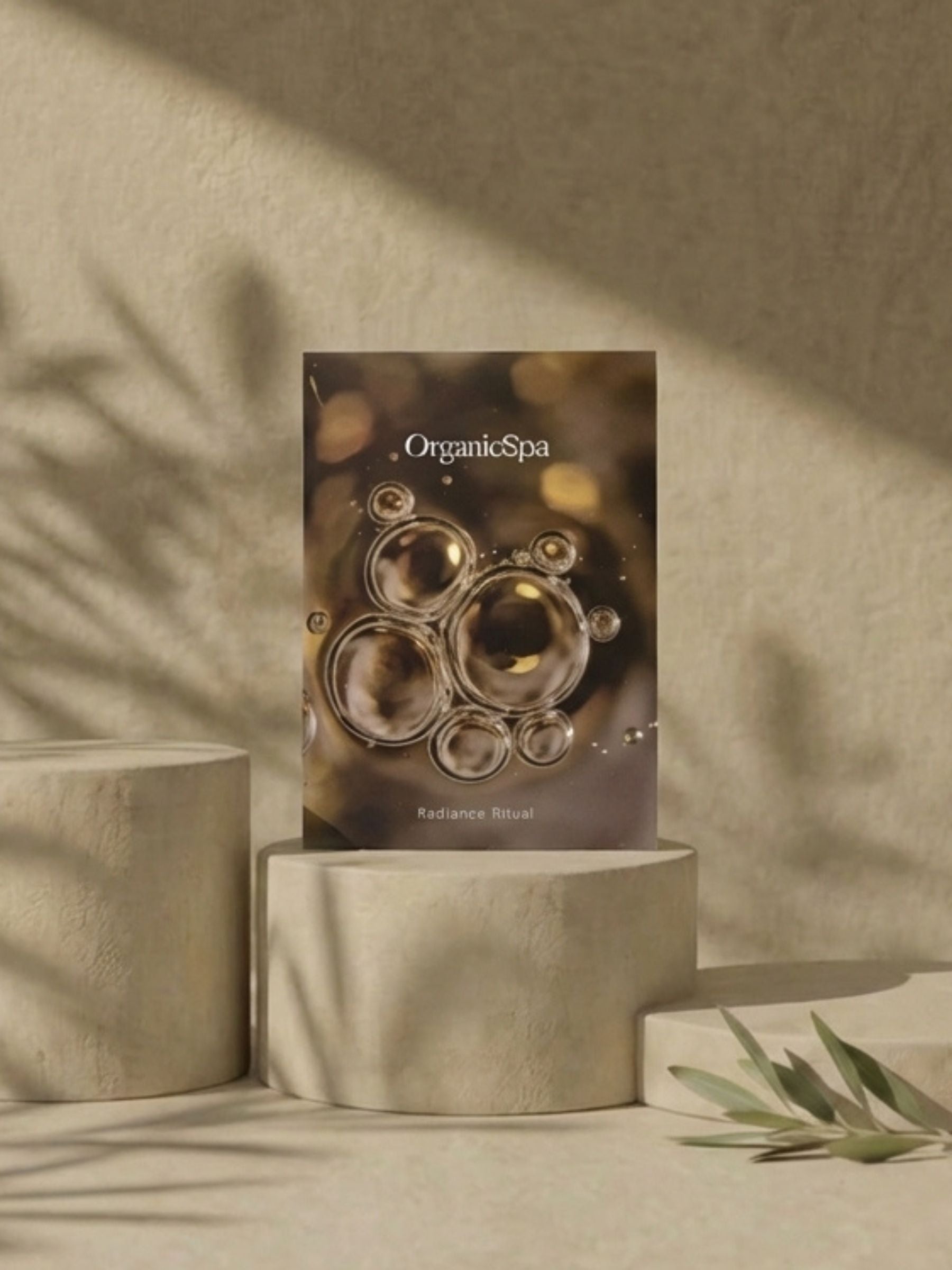 OrganicSpa Radiance Ritual box standing on stone cylindrical plinths with dramatic olive branch shadow play across a warm sandy textured wall