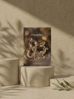 OrganicSpa Radiance Ritual box standing on stone cylindrical plinths with dramatic olive branch shadow play across a warm sandy textured wall