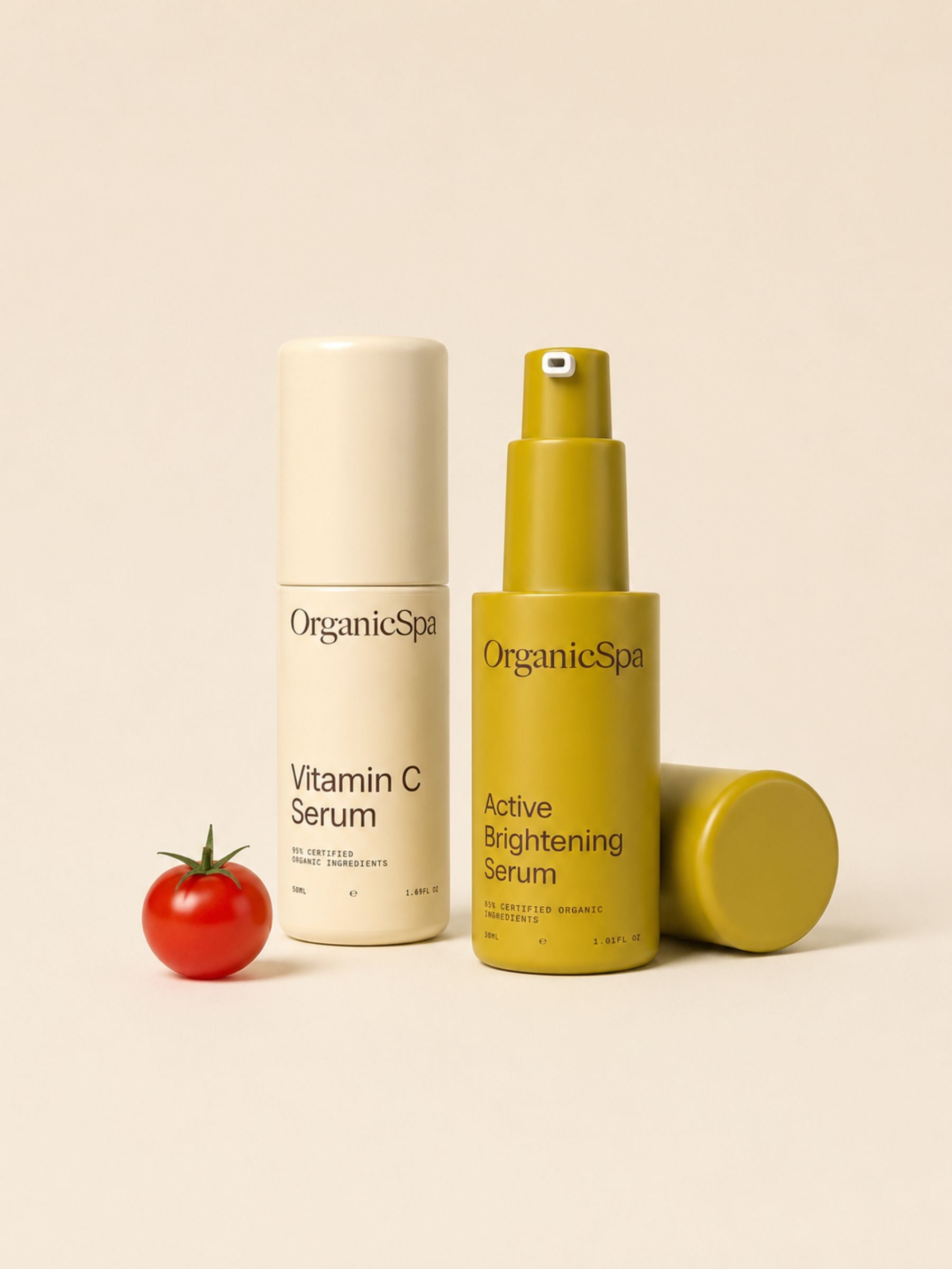 OrganicSpa Vitamin C Serum and Active Brightening Serum on a warm cream background, styled with a fresh cherry tomato. Certified Organic Skincare.