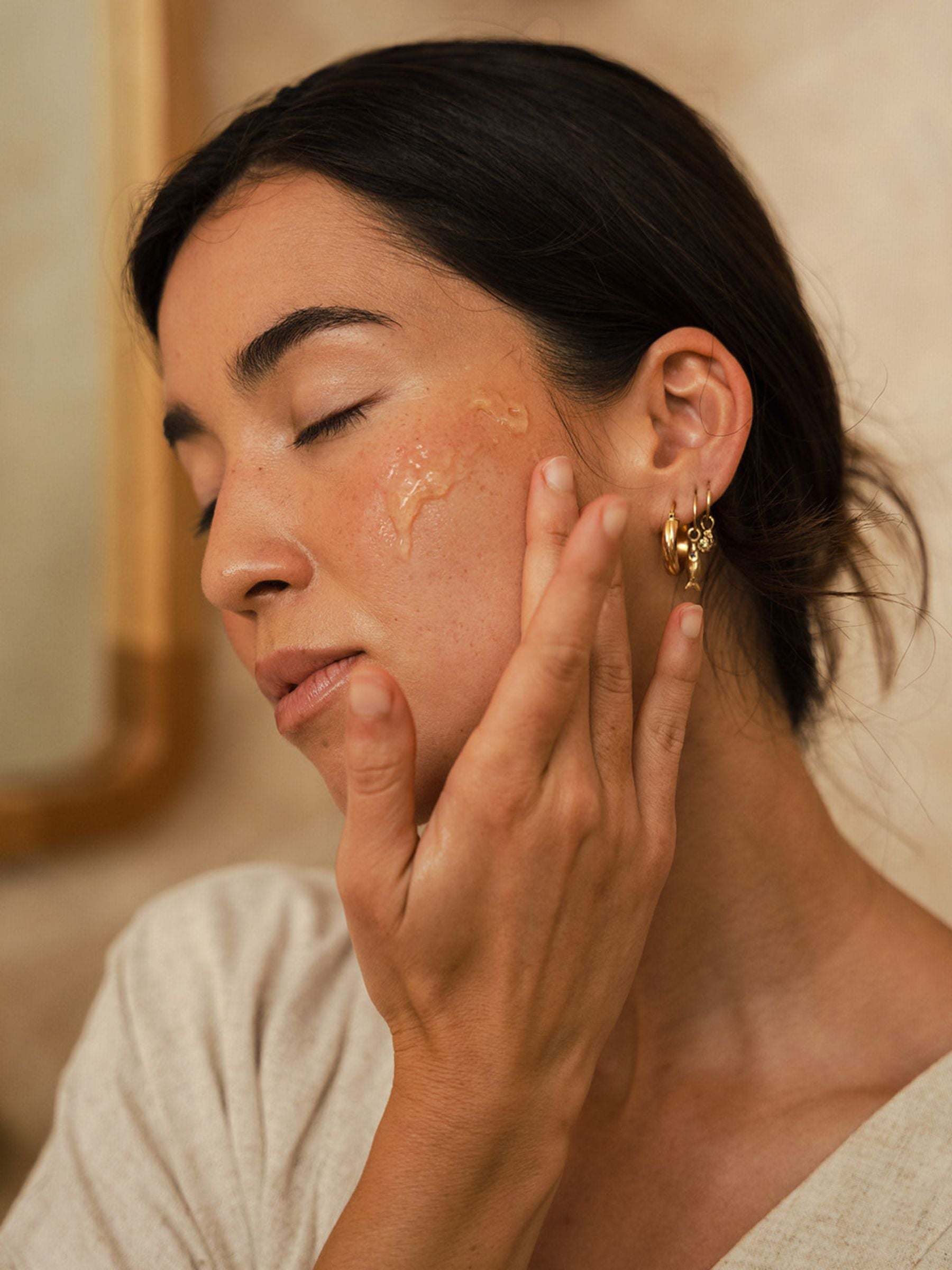 A woman gently applies OrganicSpa serum to her cheek, eyes closed, wearing a soft linen top and gold earrings. Warm, natural light. Certified Organic Skincare.