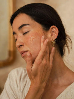 A woman gently applies OrganicSpa serum to her cheek, eyes closed, wearing a soft linen top and gold earrings. Warm, natural light. Certified Organic Skincare.