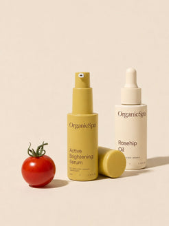 OrganicSpa Active Brightening Serum and Rosehip Oil in olive and cream bottles beside a fresh cherry tomato on a soft beige background.