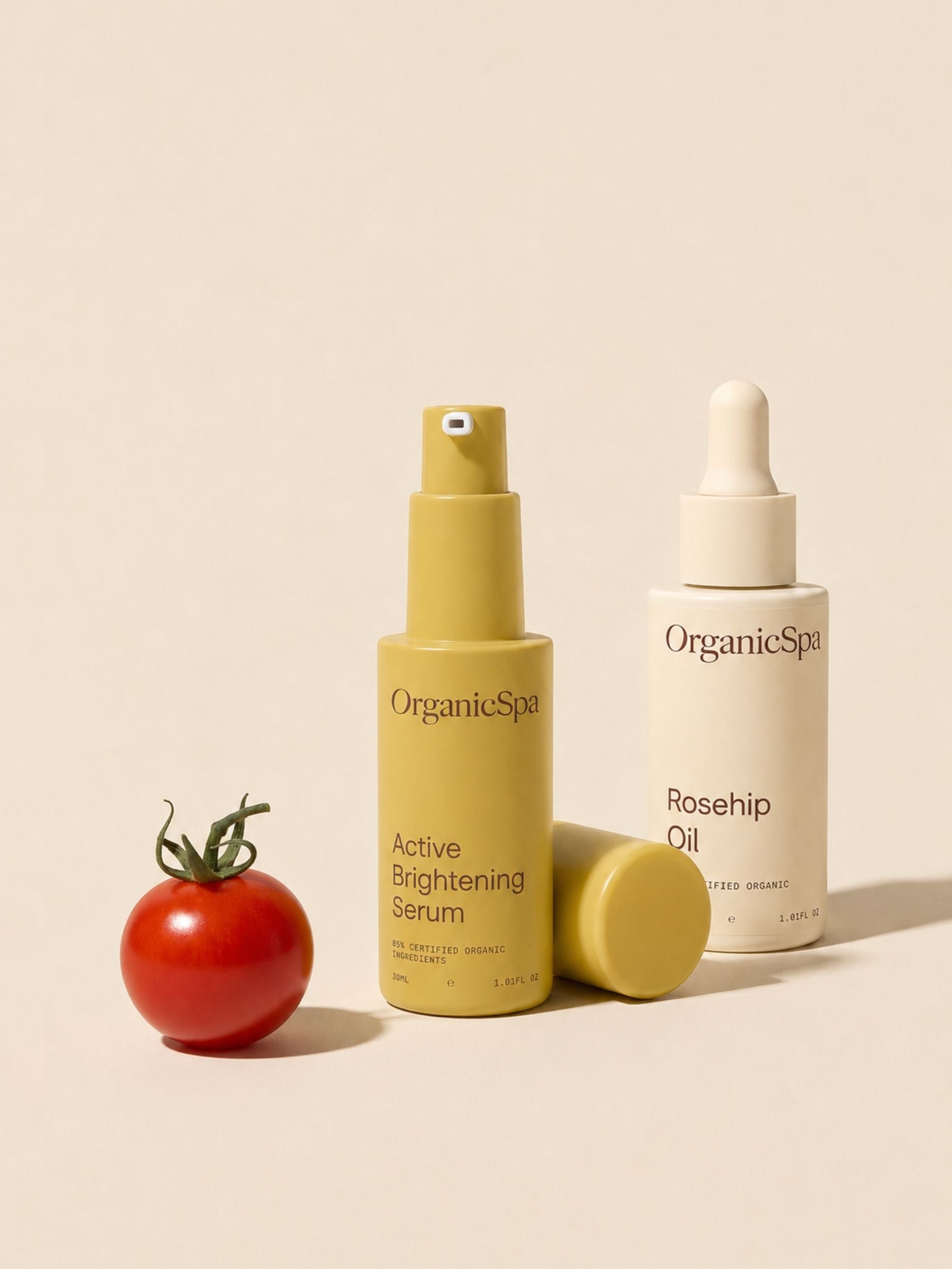 OrganicSpa Active Brightening Serum and Rosehip Oil in olive and cream bottles beside a fresh cherry tomato on a soft beige background.