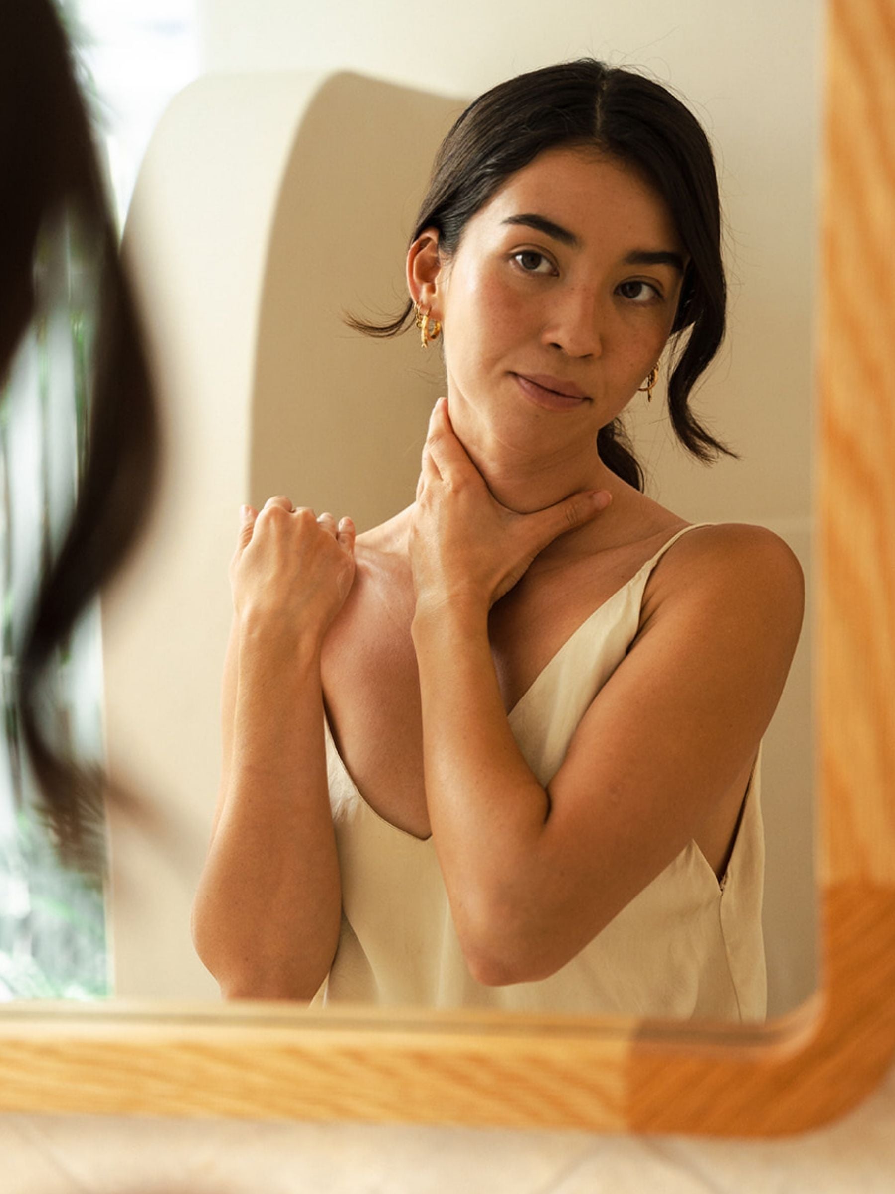 A woman in a cream slip top gazes at her reflection in a wooden-framed mirror, one hand resting on her neck, in a warm, naturally lit room.