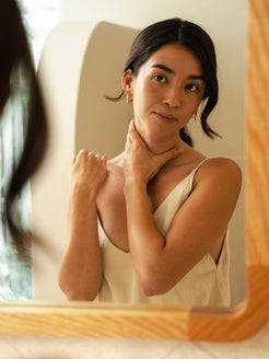 A woman in a cream slip top gazes at her reflection in a wooden-framed mirror, one hand resting on her neck, in a warm, naturally lit room.