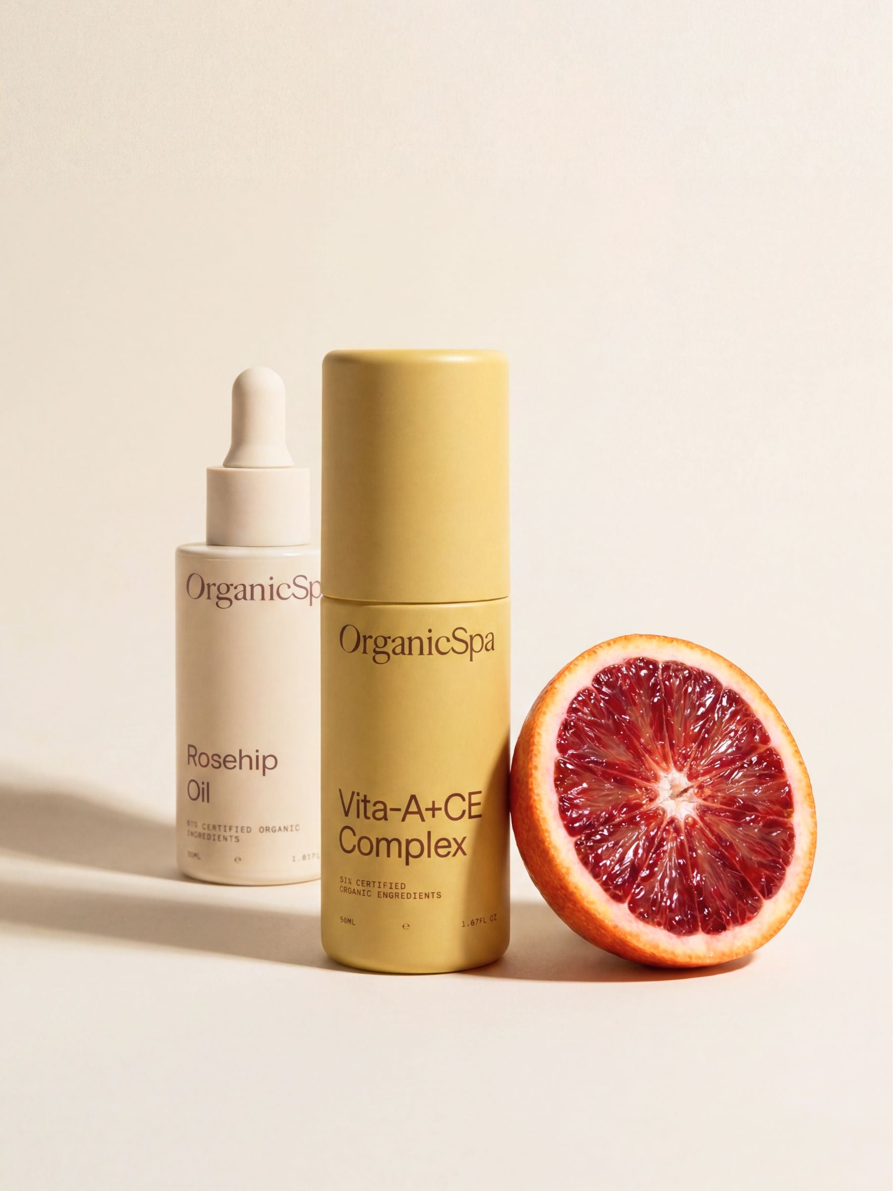 OrganicSpa Rosehip Oil and Vita-A+CE Complex in cream and olive bottles beside a halved blood orange on a soft beige background.