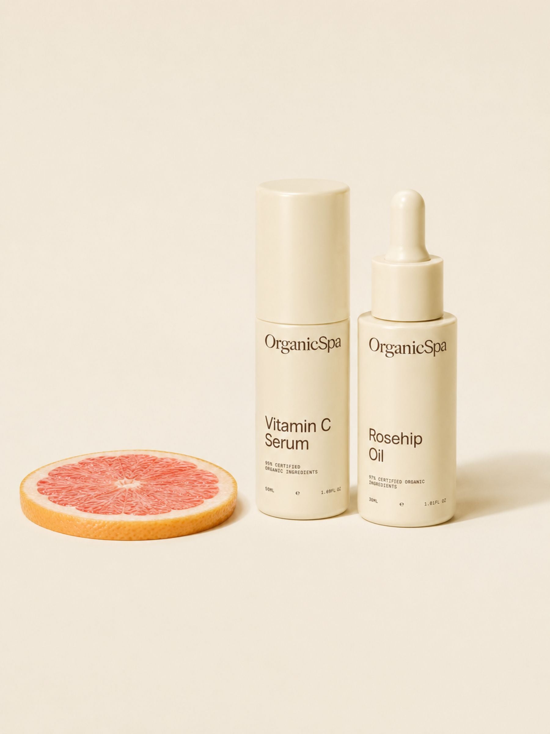 OrganicSpa Vitamin C Serum and Rosehip Oil in cream bottles beside a slice of pink grapefruit on a soft beige background.