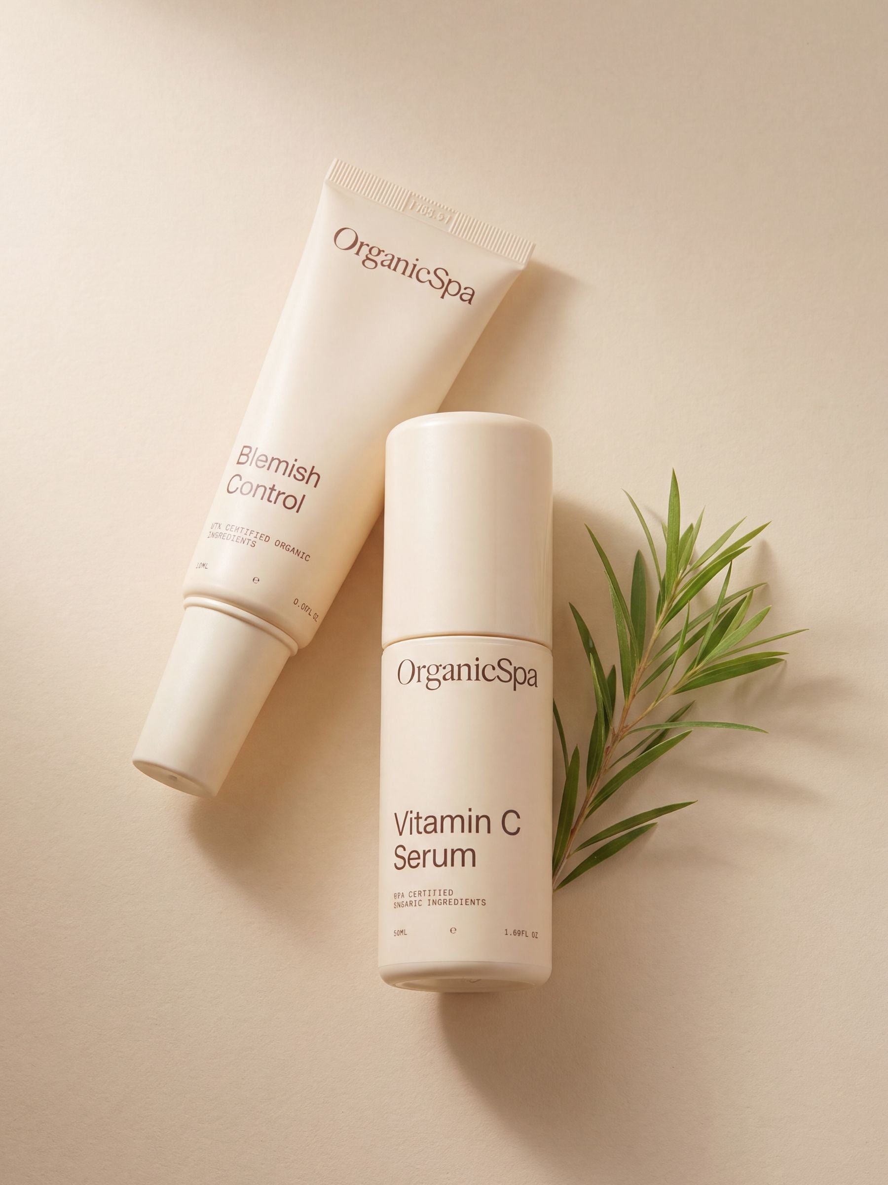 OrganicSpa Blemish Control and Vitamin C Serum laid flat on a warm cream background, styled with a fresh sprig of tea tree — a nod to the certified organic botanicals within. Certified Organic Skincare.
