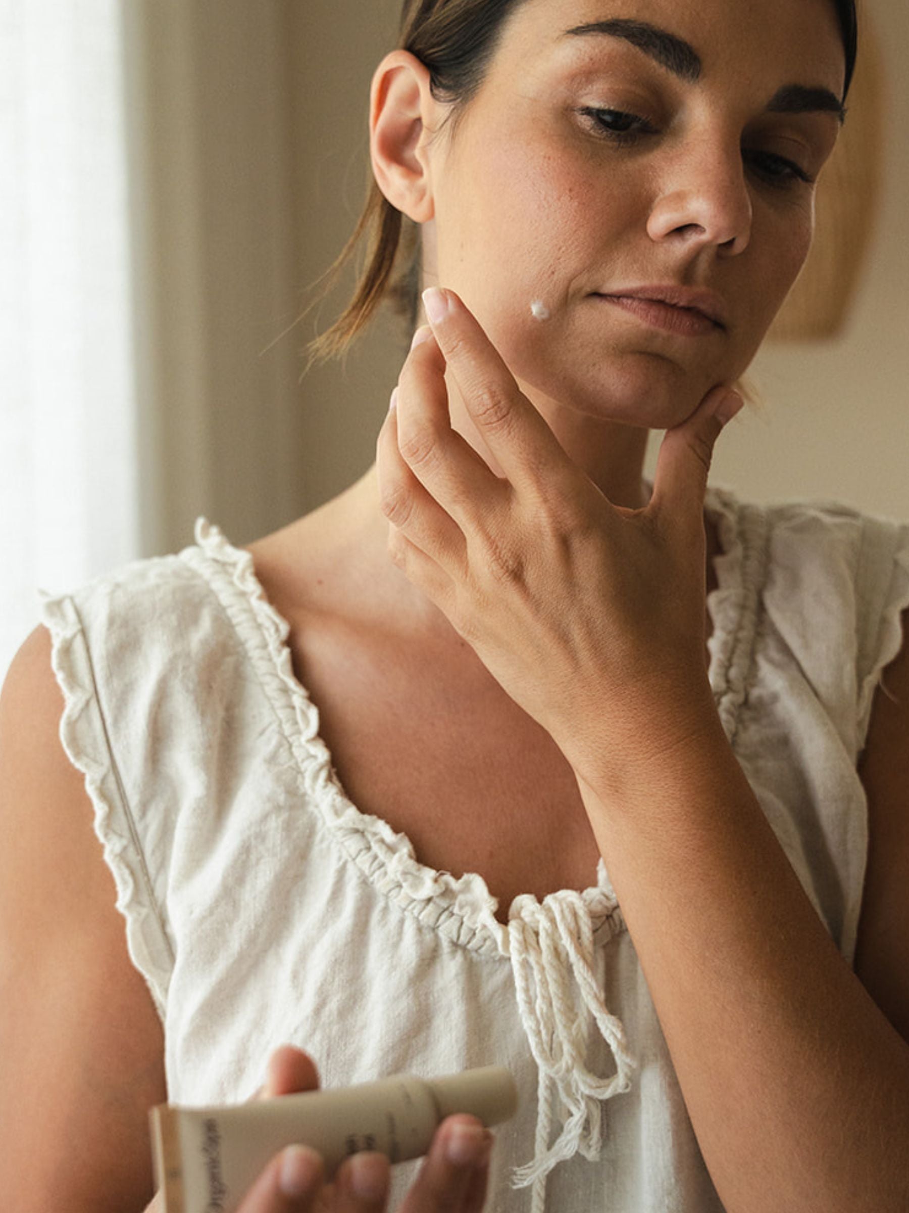 A woman in a soft white linen top applies OrganicSpa Blemish Control to her chin with a single fingertip, holding the tube in her other hand. Warm natural light, calm and considered. Certified Organic Skincare.