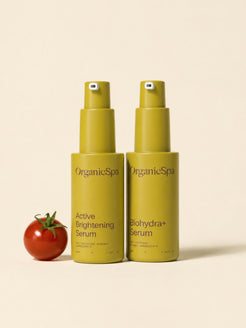 OrganicSpa Active Brightening Serum and Biohydra+ Serum in olive bottles beside a ripe cherry tomato on a soft beige background.