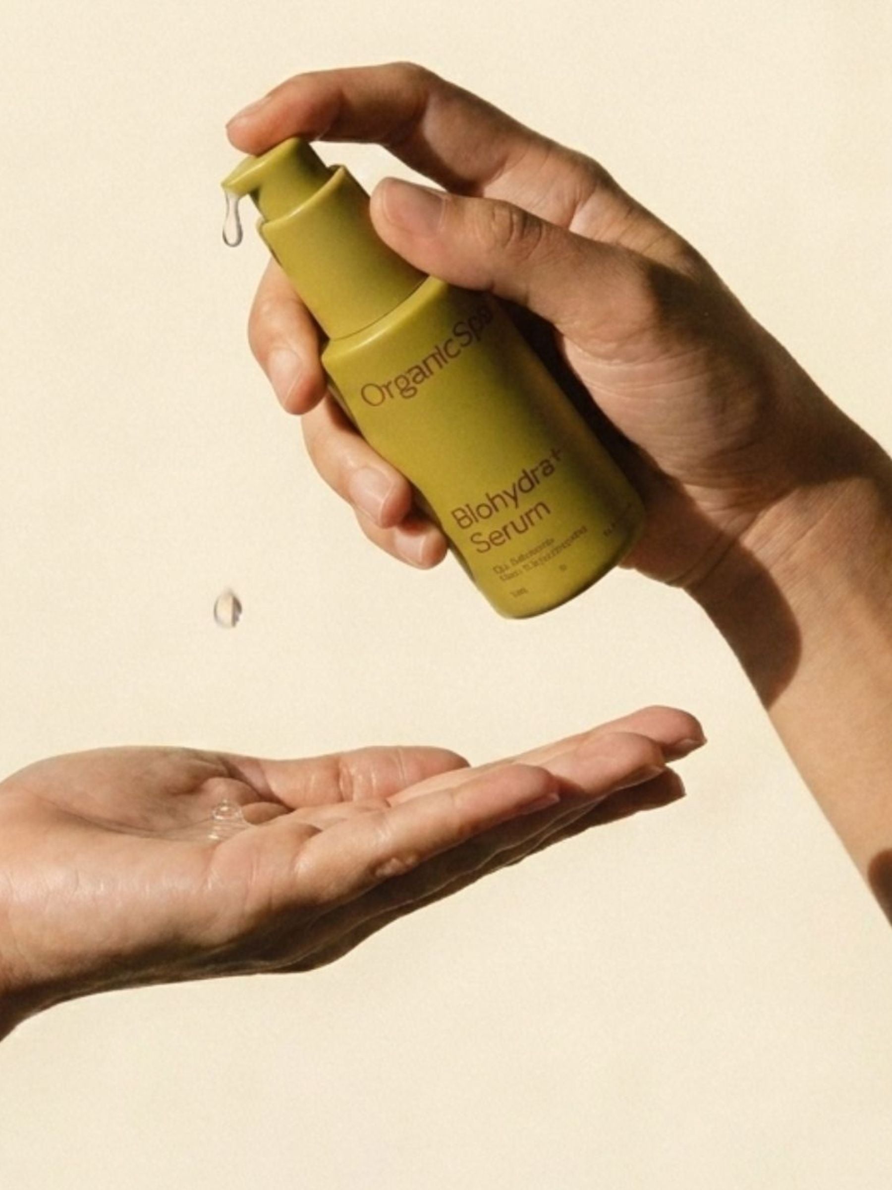 A clear drop of OrganicSpa Biohydra+ Serum dispensed from its olive bottle into an open palm in warm natural light.