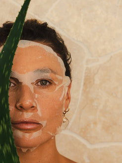 A woman wearing the OrganicSpa Hyaluronic Hydra+ Sheet Mask, photographed against a warm stone background with an aloe vera plant — showing the mask applied to the skin