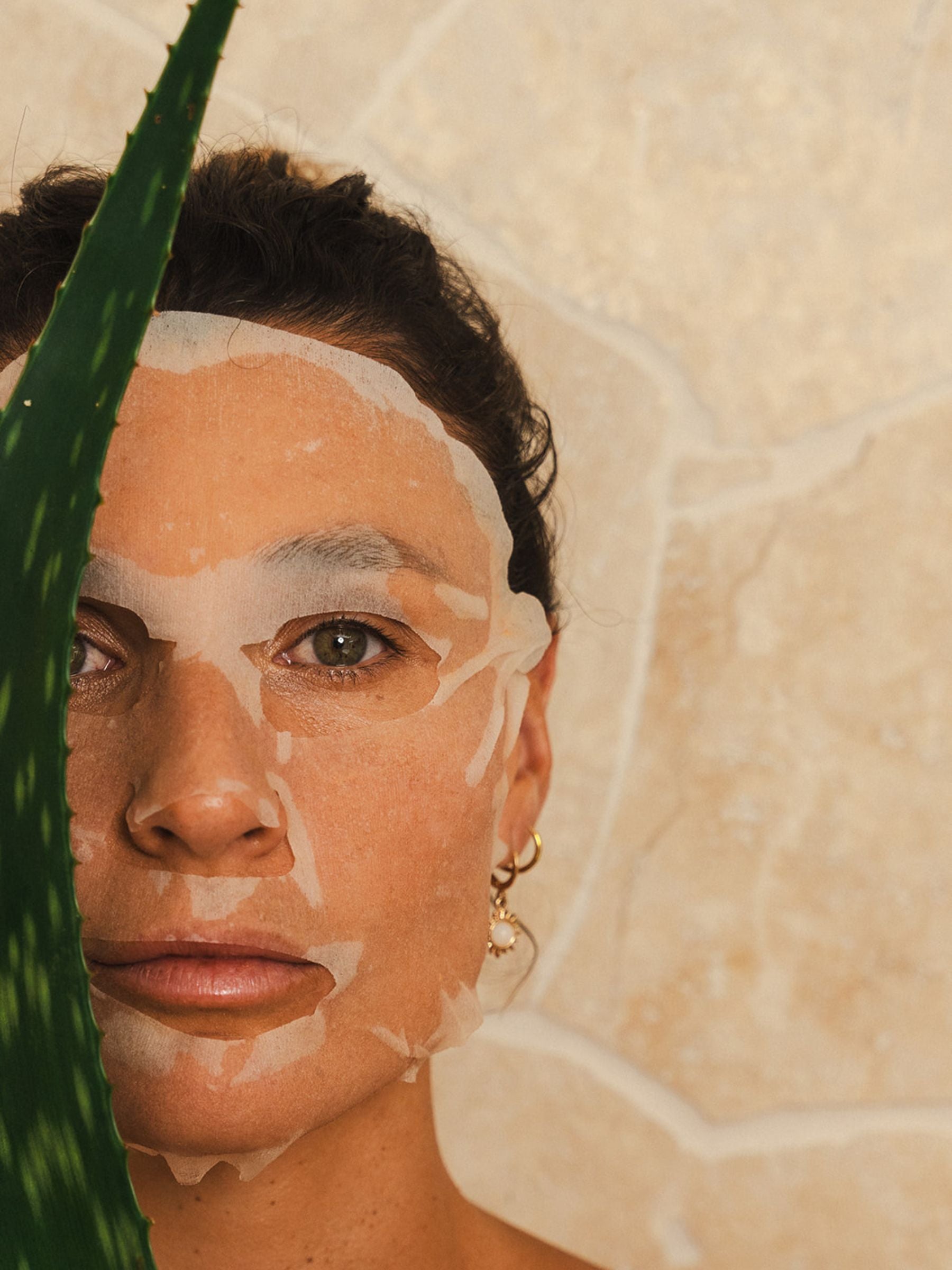 A woman wearing the OrganicSpa Hyaluronic Hydra+ Sheet Mask, photographed against a warm stone background with an aloe vera plant — showing the mask applied to the skin