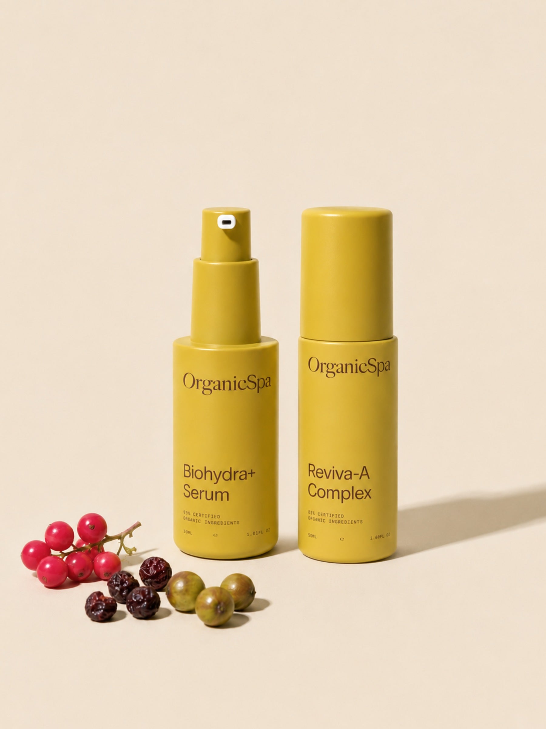 OrganicSpa Biohydra+ Serum and Reviva-A Complex on a warm cream background, styled with a cluster of native berries — including what appears to be lilly pilly and Davidson plum — nodding to the antioxidant-rich Australian botanicals within. Certified Organic Skincare.