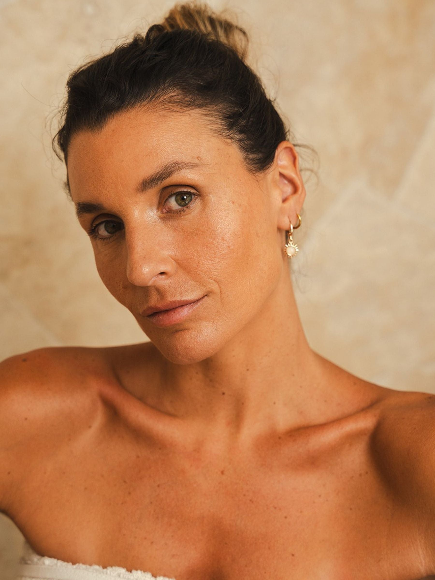 A close portrait of a woman looking directly at camera, skin dewy and makeup-free, hair loosely pinned up. Sun charm earring, warm textured background. Certified Organic Skincare.