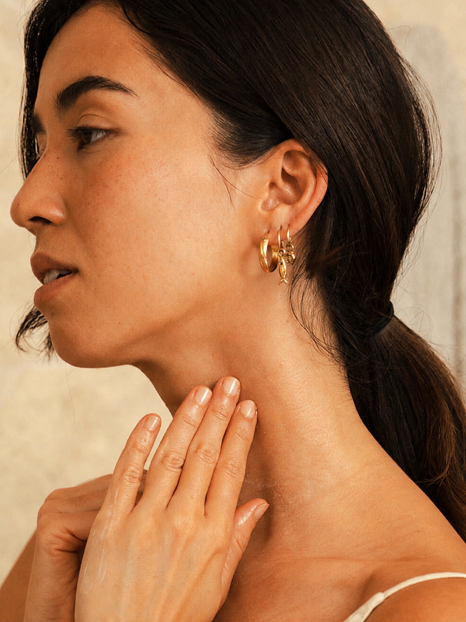 A woman in profile gently presses product into her neck and jawline with both hands, skin visibly dewy. Gold hoop earrings and a soft textured wall background. Certified Organic Skincare.