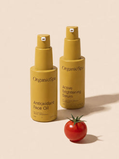OrganicSpa Antioxidant Face Oil and Active Brightening Serum on a warm cream background, styled with a fresh cherry tomato — a natural source of lycopene and antioxidants that reflects the certified organic ingredients within. Certified Organic Skincare.