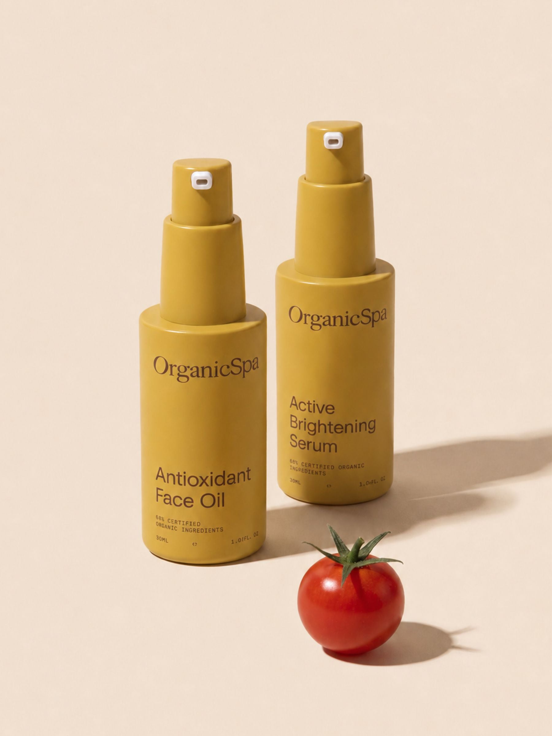 OrganicSpa Antioxidant Face Oil and Active Brightening Serum on a warm cream background, styled with a fresh cherry tomato — a natural source of lycopene and antioxidants that reflects the certified organic ingredients within. Certified Organic Skincare.