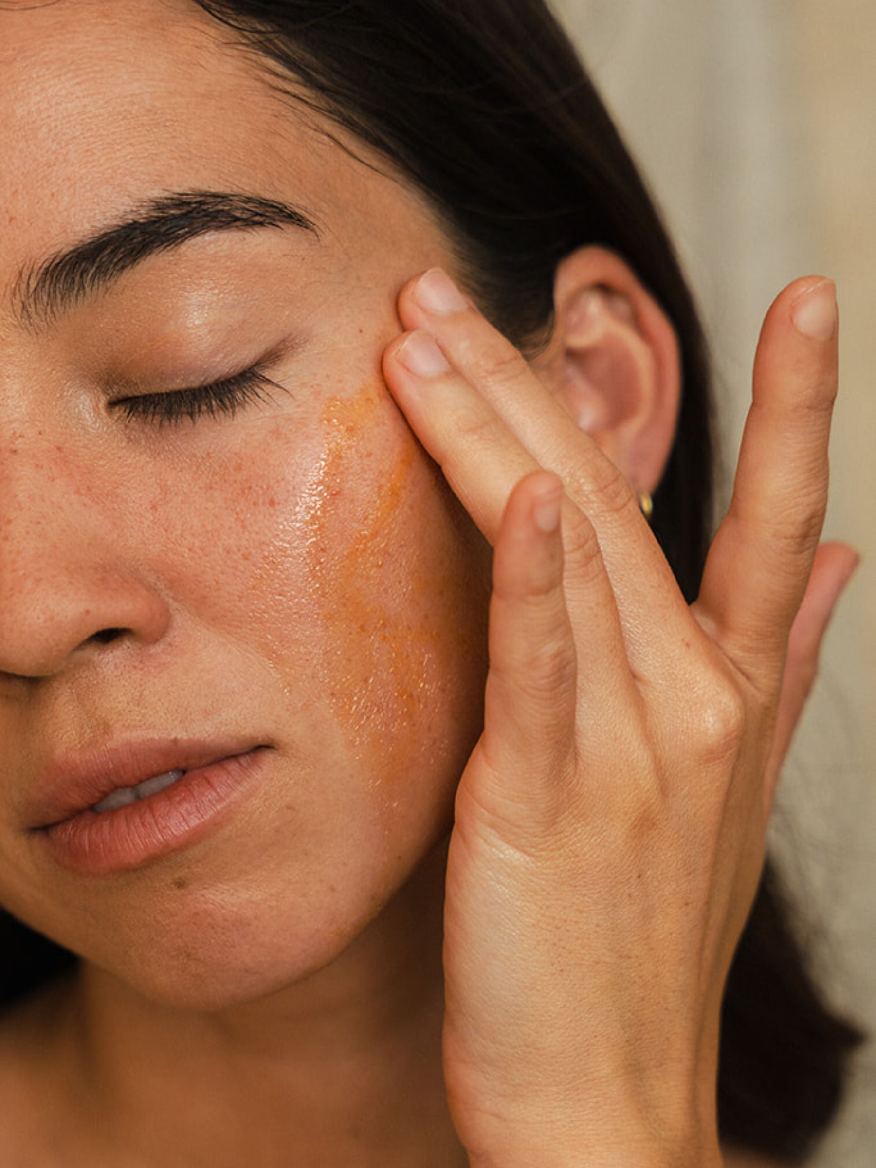 A close-up of a woman gently pressing OrganicSpa Antioxidant Face Oil into her cheek, eyes closed. The rich amber-toned oil is visible on skin against a warm neutral background. Certified Organic Skincare.
