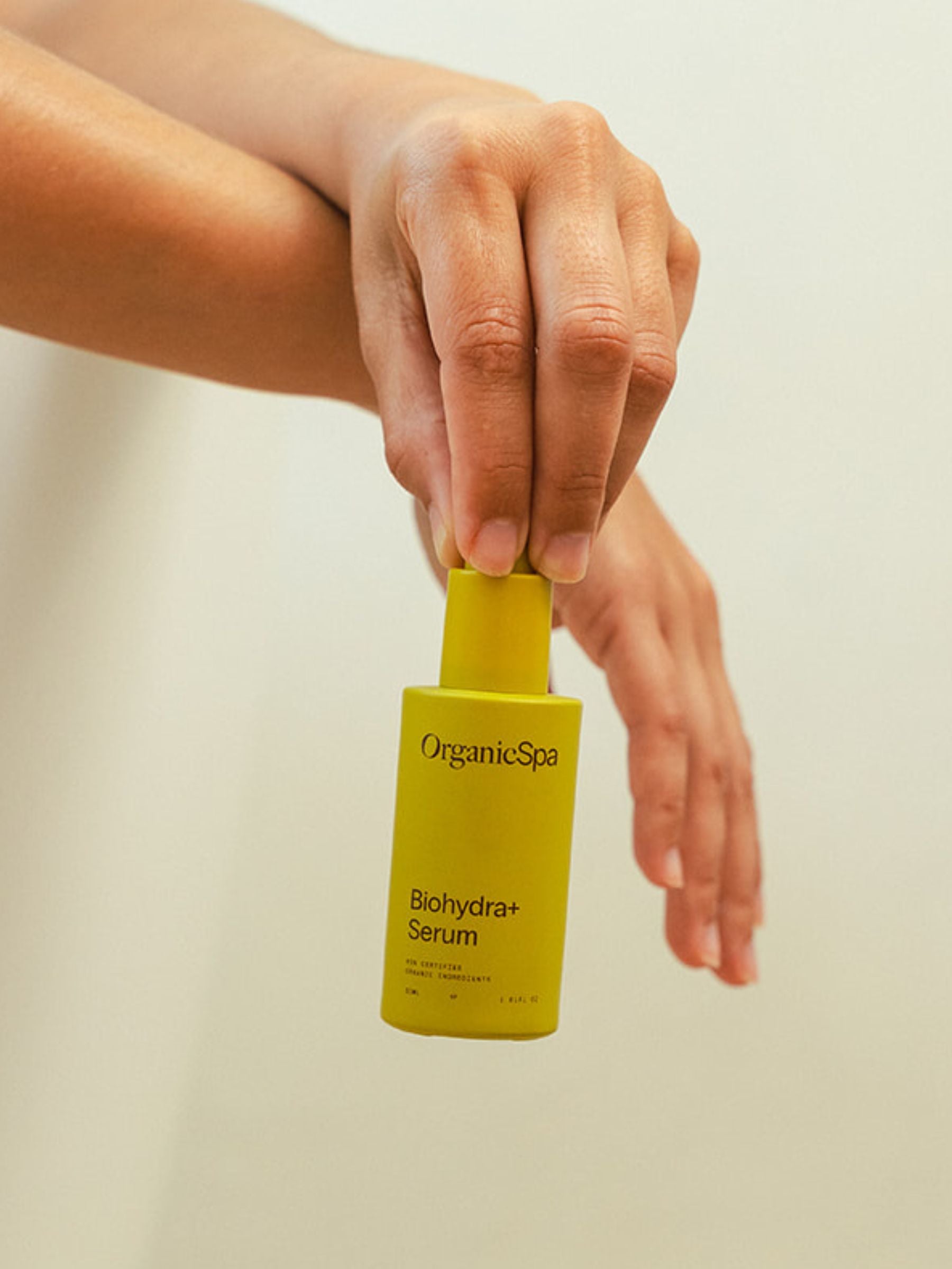 A hand holds the OrganicSpa Biohydra+ Serum upright against a soft white background, with a second hand resting gently below. Certified Organic Skincare.