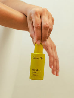 A hand holds the OrganicSpa Biohydra+ Serum upright against a soft white background, with a second hand resting gently below. Certified Organic Skincare.