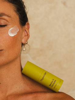 A close-up of a woman with a dollop of OrganicSpa Reviva-A Complex on her cheekbone, eyes closed, with the product bottle resting against her bare shoulder. Sun charm earring and warm sandy background. Certified Organic Skincare.