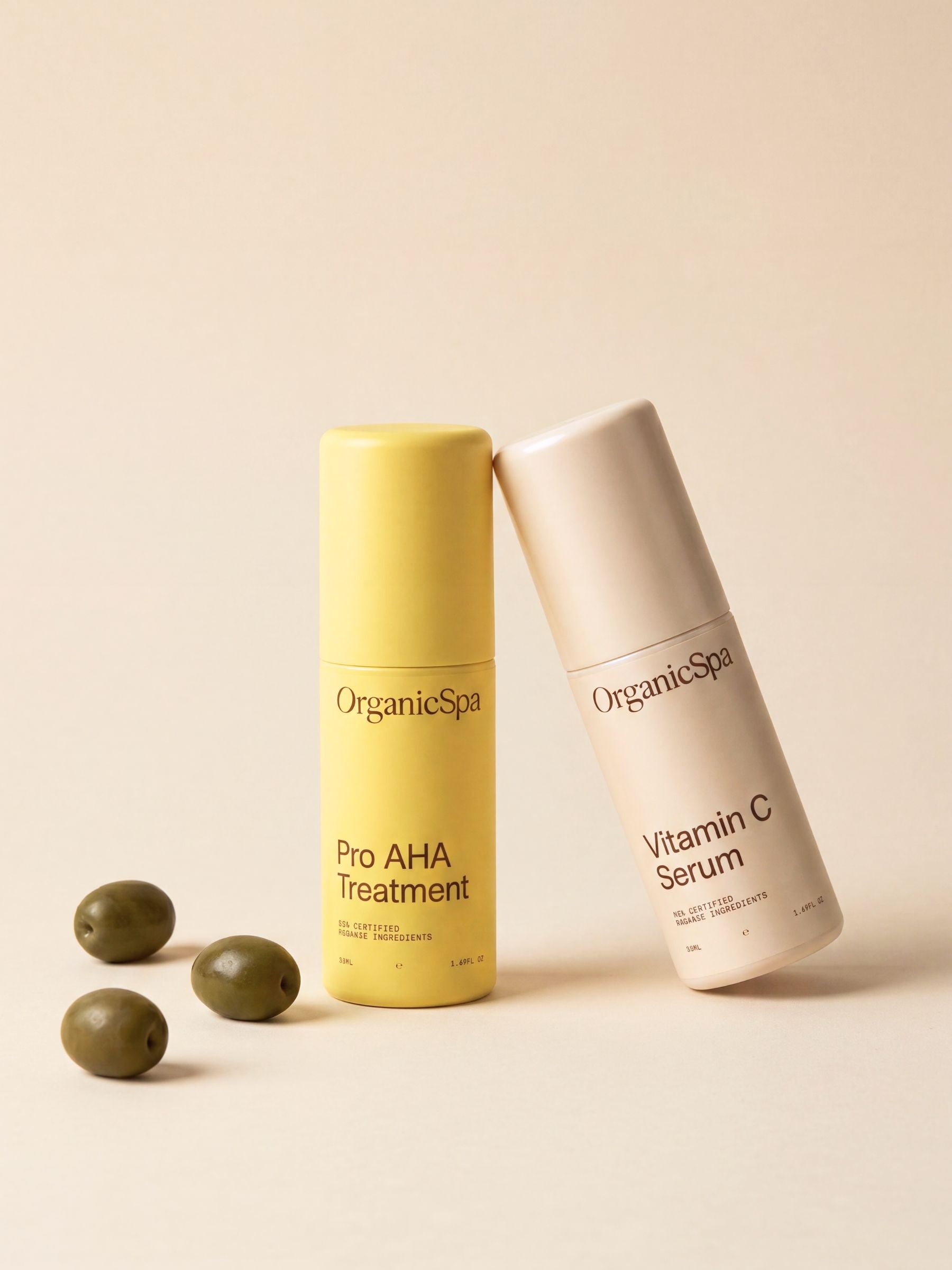 OrganicSpa Pro AHA Treatment and Vitamin C Serum on a warm cream background, styled with fresh green olives — a nod to the olive-derived organic ingredients within. Certified Organic Skincare.