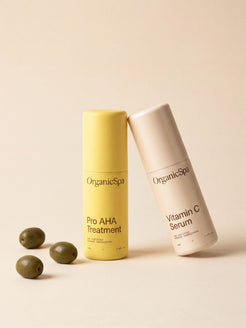 OrganicSpa Pro AHA Treatment and Vitamin C Serum on a warm cream background, styled with fresh green olives — a nod to the olive-derived organic ingredients within. Certified Organic Skincare.