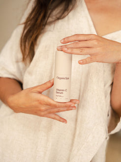 A woman in a soft linen top cradles the OrganicSpa Vitamin C Serum between both hands against a warm neutral background. Certified Organic Skincare.