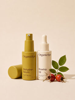 OrganicSpa Biohydra+ Serum and Rosehip Oil on a warm cream background, styled with fresh rosehip berries and leaves — the hero certified organic ingredient in the oil. Certified Organic Skincare.