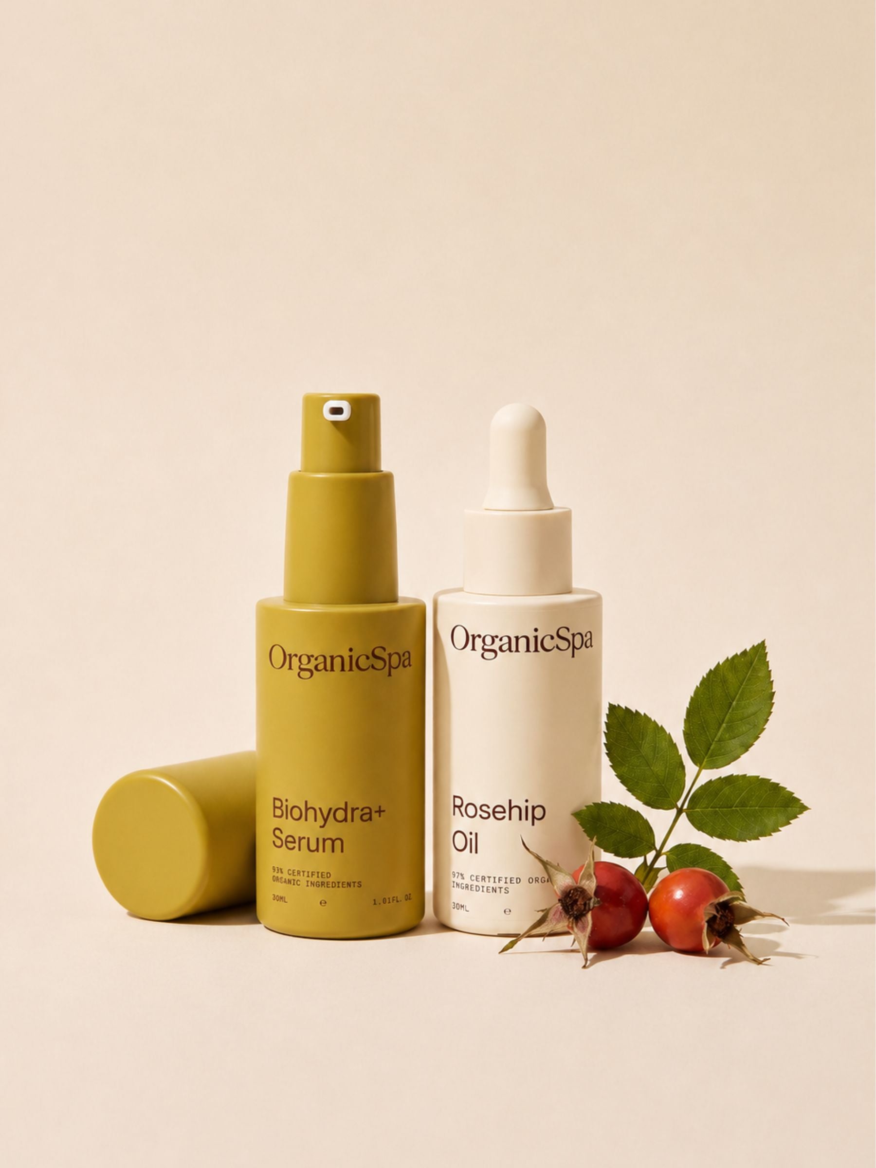 OrganicSpa Biohydra+ Serum and Rosehip Oil on a warm cream background, styled with fresh rosehip berries and leaves — the hero certified organic ingredient in the oil. Certified Organic Skincare.