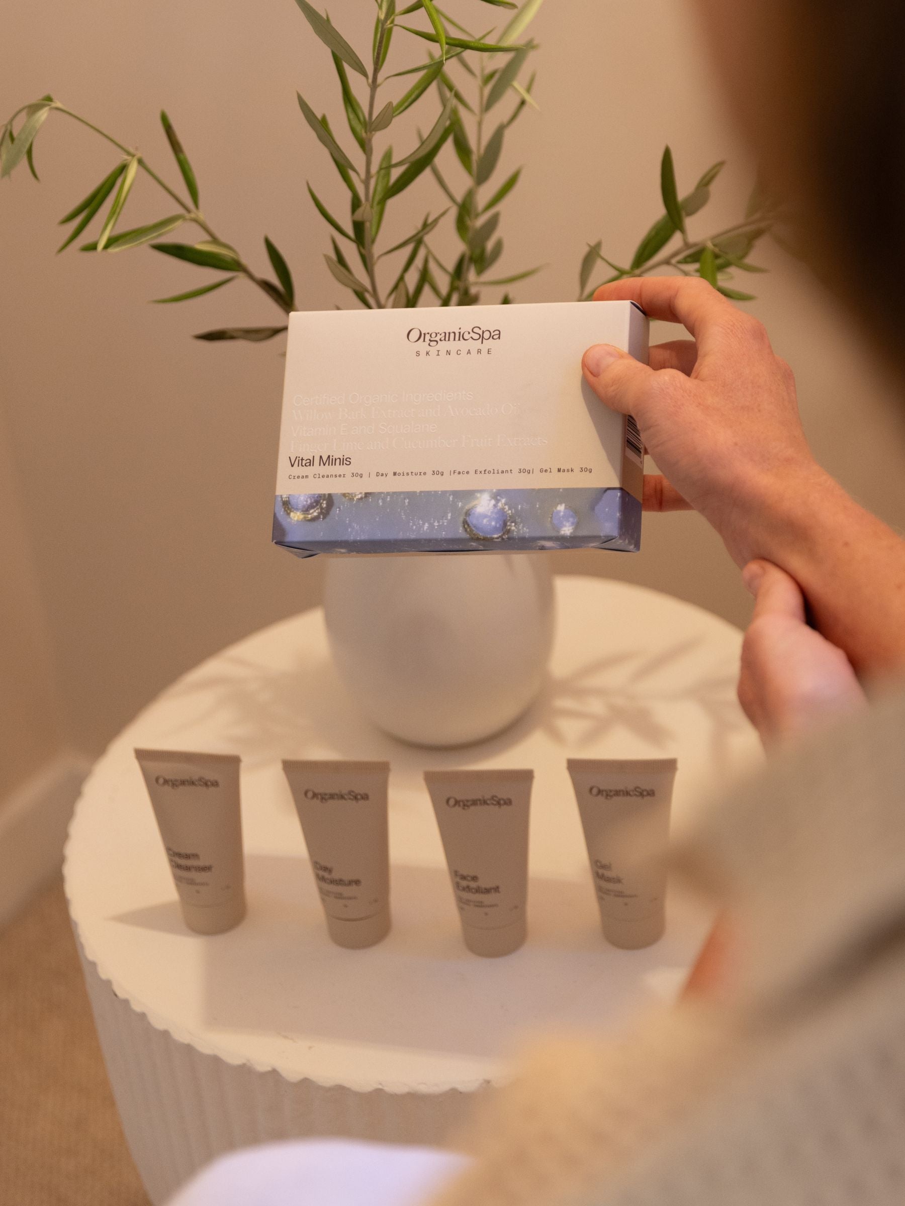 Hand holding OrganicSpa Vital Minis box above the four 30g travel-sized tubes — Cream Cleanser, Day Moisture, Face Exfoliant and Gel Mask — Certified Organic skincare for normal and combination skin.