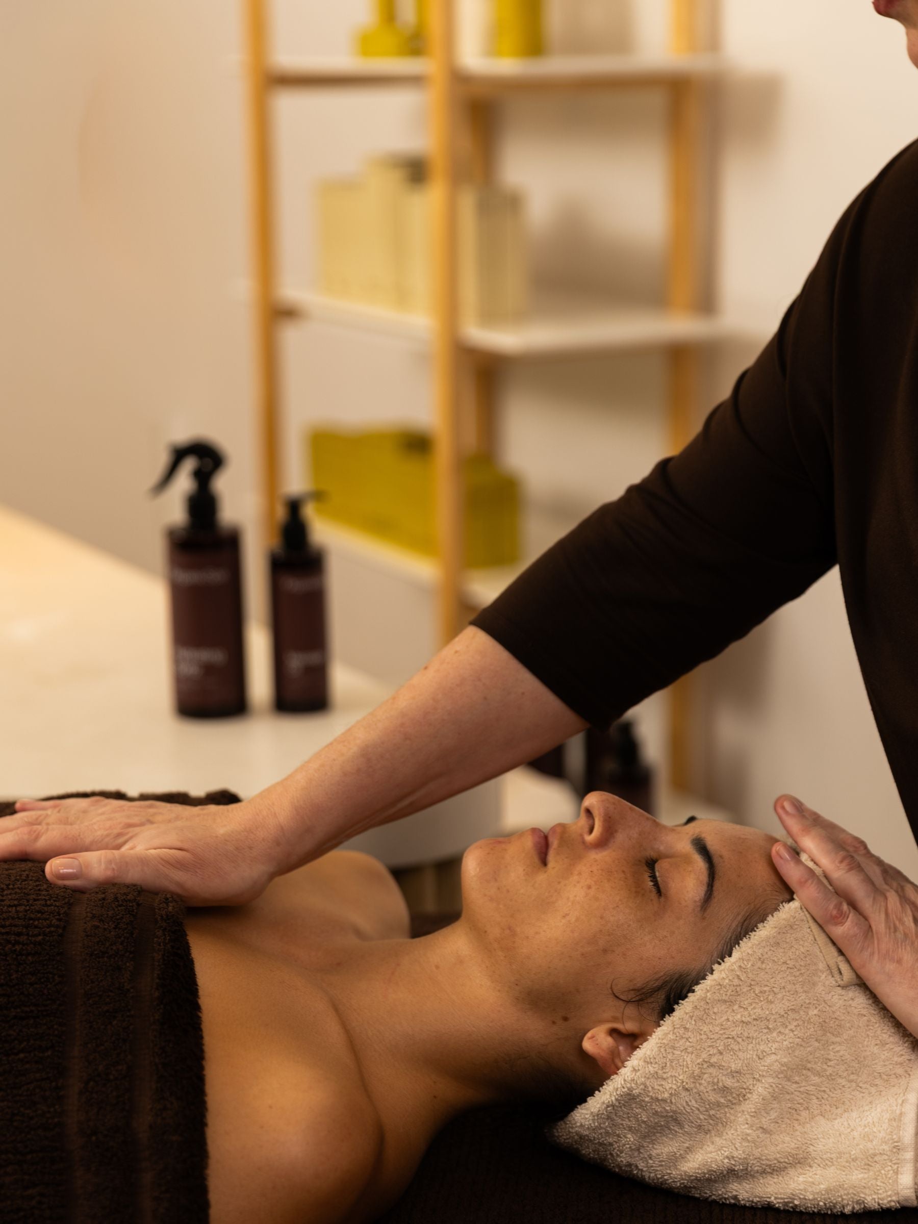 A skin therapist performing a facial massage, hands cradling a client's head and décolletage. OrganicSpa products are visible on the treatment bench behind them.