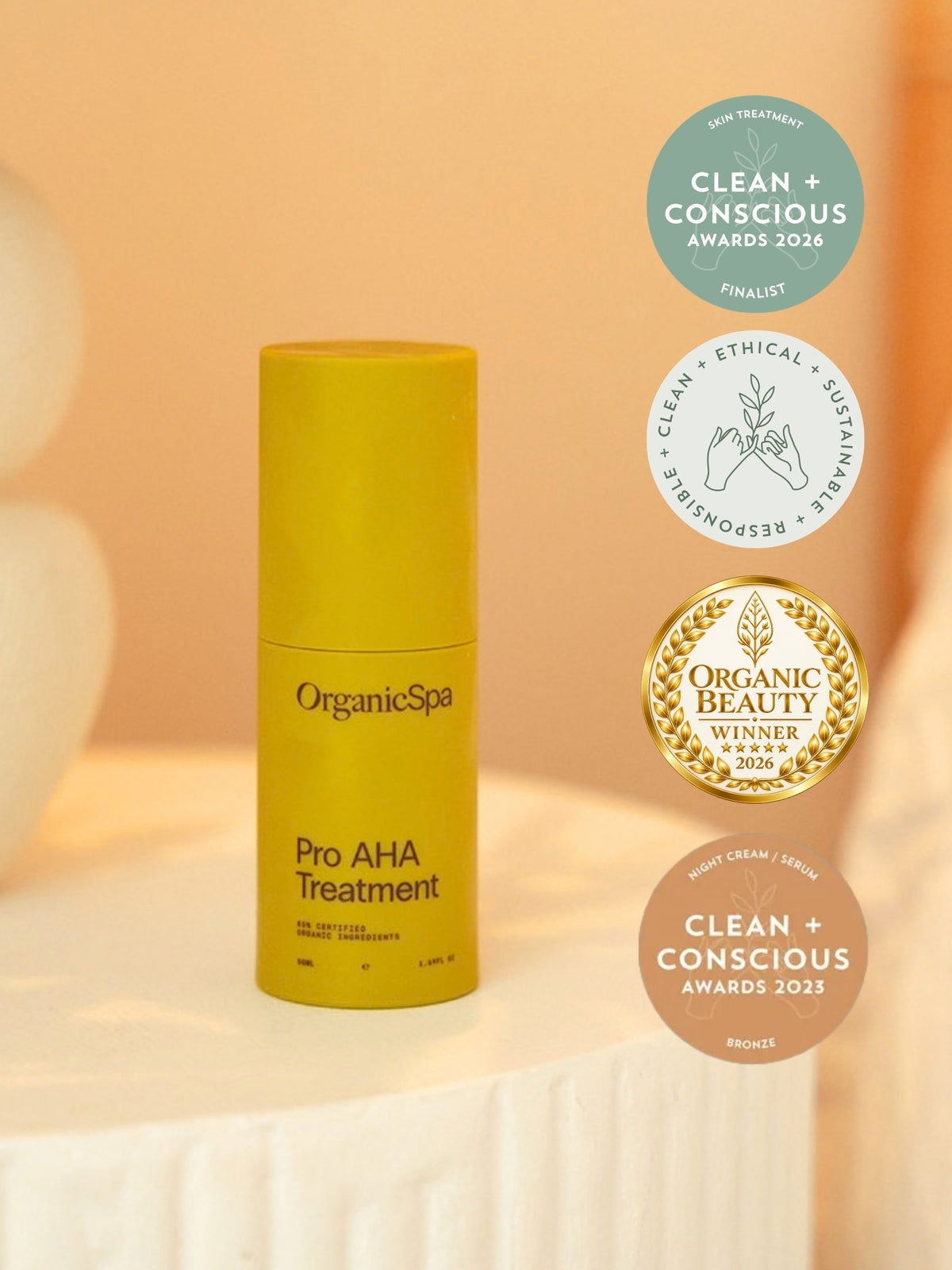 OrganicSpa Pro AHA Treatment, a chartreuse bottle on a cream plinth. Four badges beside it: finalist in the Skin Treatment category at the Clean + Conscious Awards 2026, the awards' clean, ethical, sustainable and responsible seal, Organic Beauty winner 2026, and Bronze in the Night Cream/Serum category at the Clean + Conscious Awards 2023.