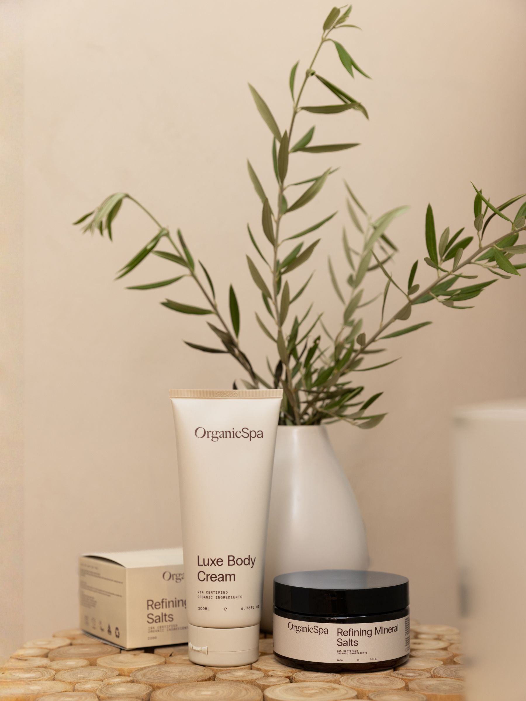 OrganicSpa Luxe Body Cream tube and Refining Mineral Salts jar displayed on a timber surface with their kraft box packaging and a white vase of green stems
