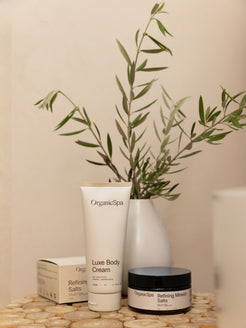 OrganicSpa Luxe Body Cream tube and Refining Mineral Salts jar displayed on a timber surface with their kraft box packaging and a white vase of green stems
