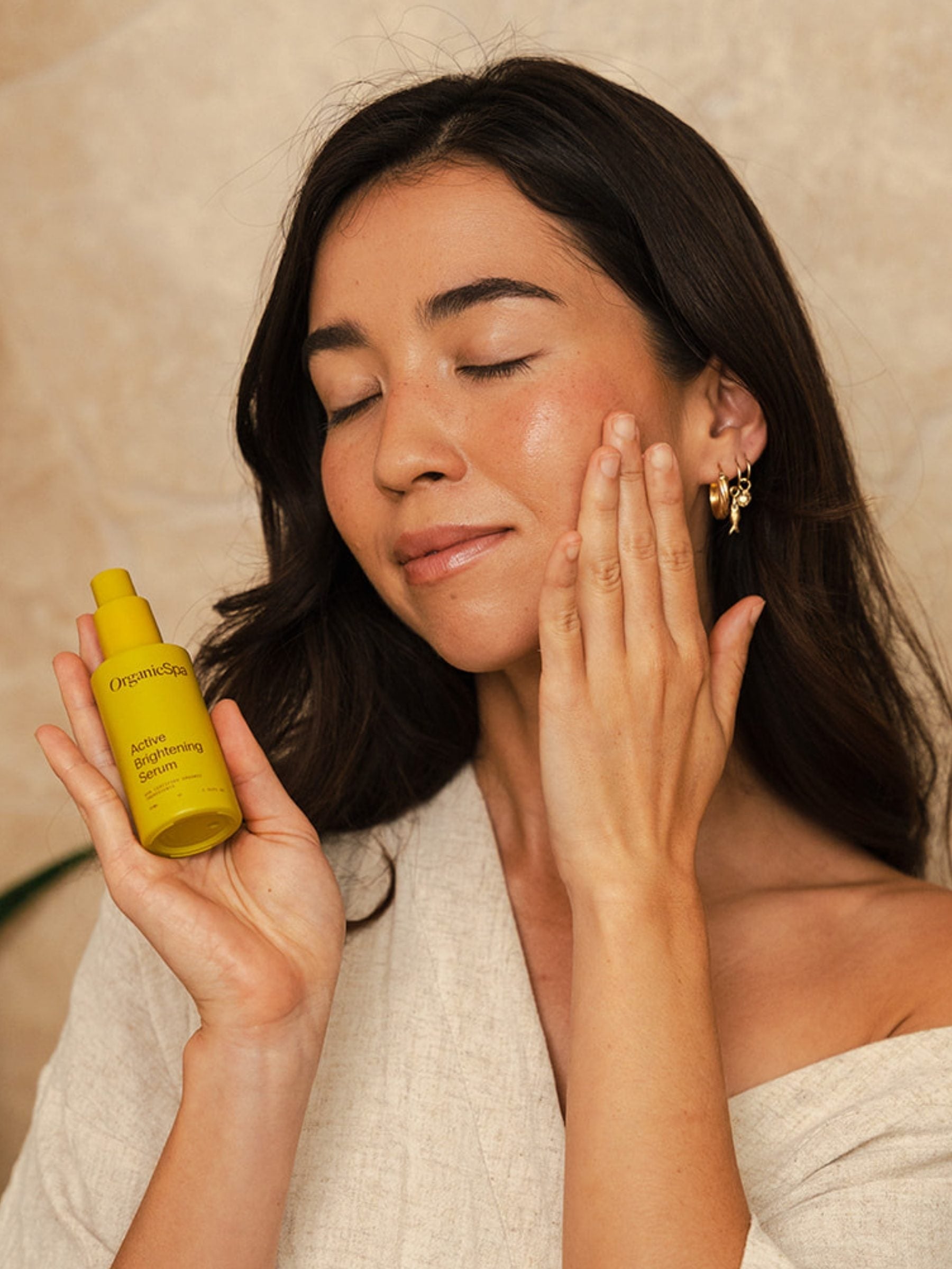 A woman holding OrganicSpa Active Brightening Serum and pressing it into her cheek with her eyes closed.