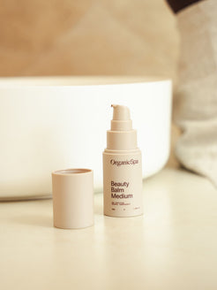 Organicspa Beauty Balm Medium bottle with lid beside it, styled on a bathroom vanity in warm natural light.